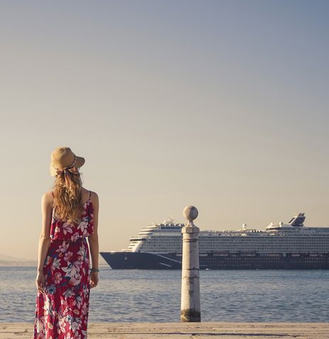 Frau mit einem bunten langen Kleid steht an einer Promenade und schaut auf ein Kreuzfahrtschiff