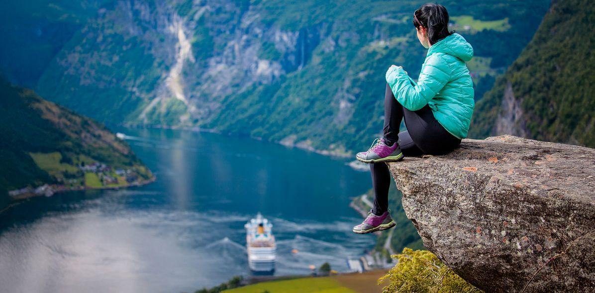 Eine Frau sitzt auf einem Felsen und genießt den Panoramablick auf den Geiranger Fjord in Norwegen, während sie ein Kreuzfahrtschiff beobachtet