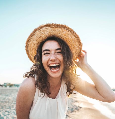 Frau mit Sonnenhut steht am Strand und lacht in die Kamera