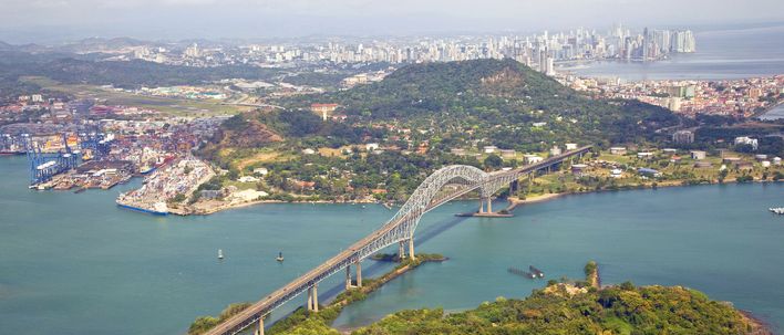 Luftaufnahme des Panamakanals mit Brücke über bewaldete Hügel und Skyline im Hintergrund