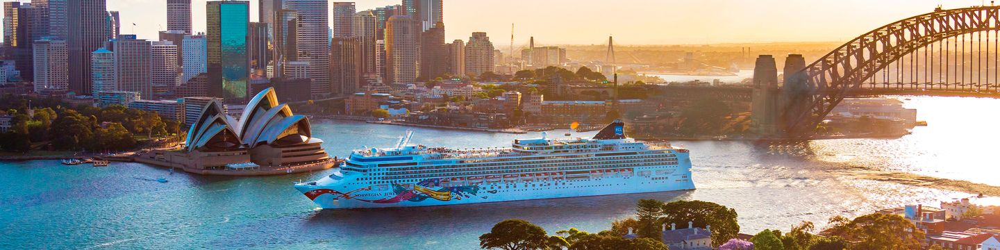 Ein Schiff der Norwegian Cruise Line fährt bei Sonnenuntergang zwischen Opernhaus und Harbour Bridge durch Sydney.