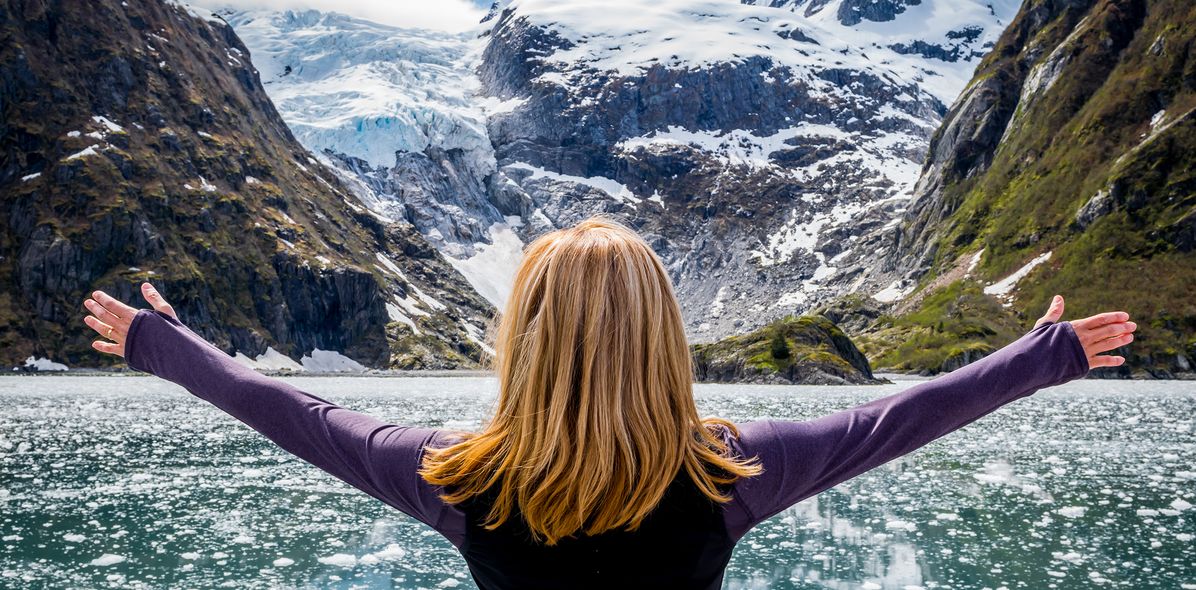 Reisende blickt mit ausgebreiteten Armen auf schneebedeckte Berge und einen Gletscher im Kenai-Fjords-Nationalpark in Alaska