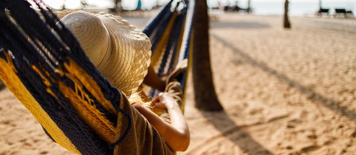 Person entspannt in einer Hängematte am sonnigen Sandstrand mit Strohhüten im Hintergrund