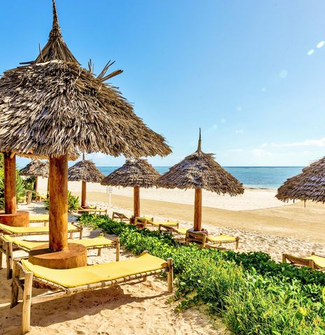 Blick aufs Meer von Sansibar über einen typischen Strand mit Sonnenschirmen und -liegen aus Palmen und Bäumen