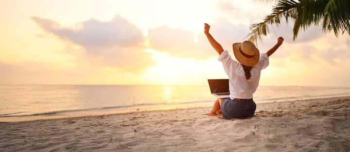 Frau sitzt mit Laptop am Strand vor dem Meer und genießt den Sonnenaufgang