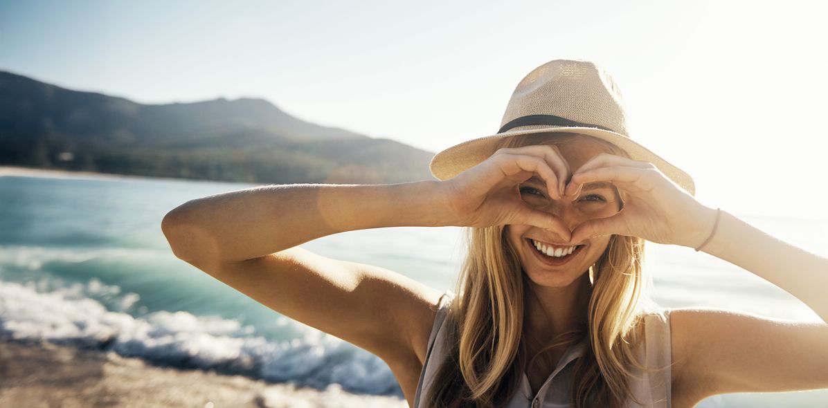 Frau mit Sommerhut am Strand formt mit den Händen ein Herz und schaut hindurch
