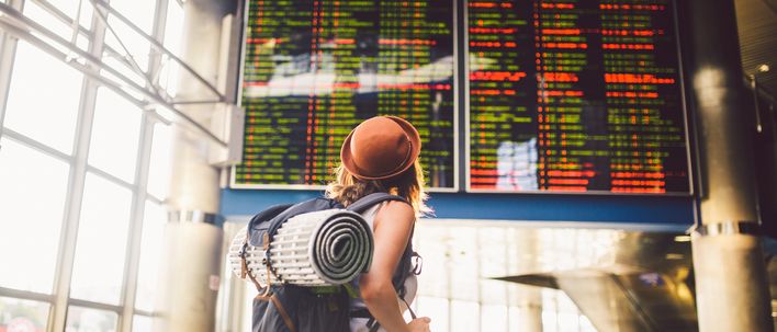 Frau mit Rucksack und Hut schaut auf die Abflugtafel im Flughafen
