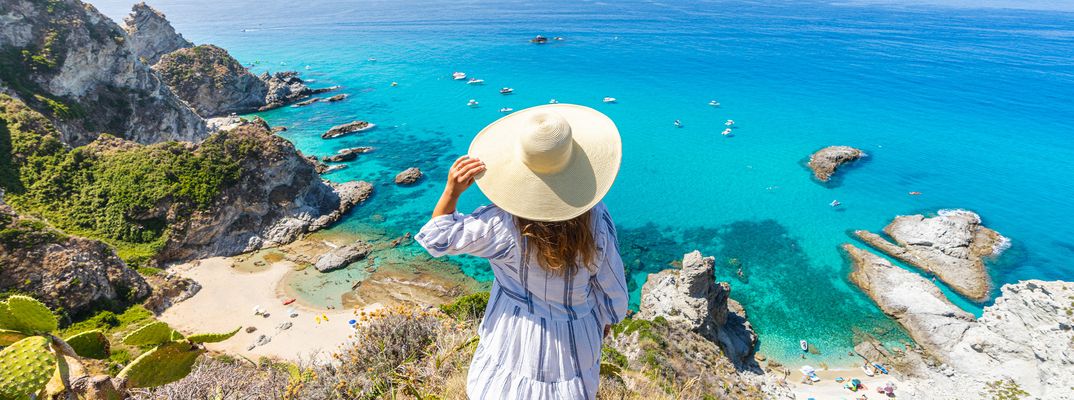 Eine Frau mit Hut steht auf Klippen und schaut auf das türkisfarbende Meer 