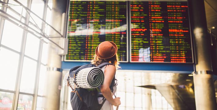 Frau mit Rucksack und Hut schaut auf die Abflugtafel im Flughafen