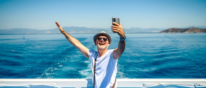 Fröhlicher Mann mit Sonnenhut macht ein Selfie an Bord eines Schiffes mit Blick auf das tiefblaue Meer