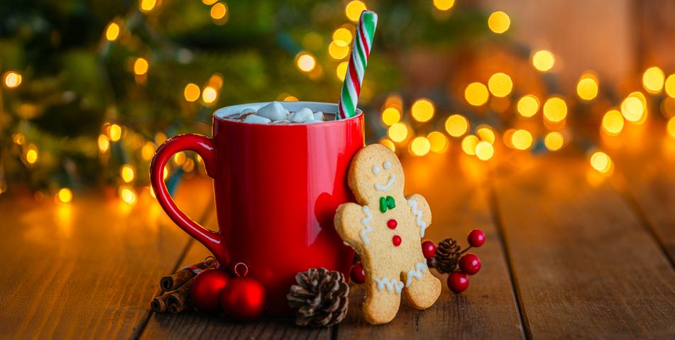 Ein Lebkuchenmännchen steht vor einer roten Weihnachtstasse mit Kakao vor einem beleuchtetem Weihnachtsbaum mit Berge&Meer Superzeichen