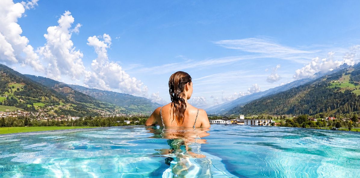Rückansicht einer Frau in einem Infinity-Pool, die auf eine weite Landschaft mit Bergen und einen wolkenbehangenen Himmel blickt