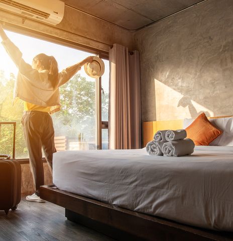Frau steht mit Koffer im Hotelzimmer am Bett und blickt durch das große Fenster ins Licht und reißt die Arme hoch