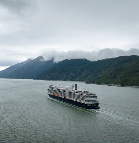 Ein Schiff der Holland America Line fährt unter grauen Wolken durch einen weiten Fjord zwischen grünen Bergen.