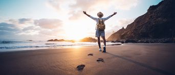 Reisender mit Rucksack genießt den Sonnenuntergang am Strand mit ausgebreiteten Armen