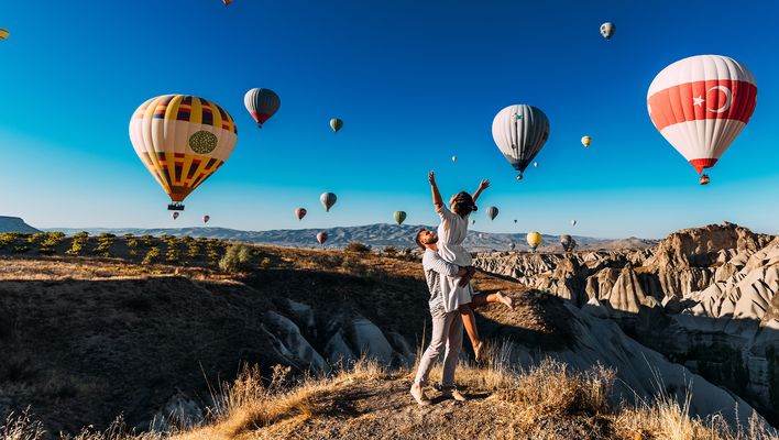 Frau mit ausgebreiteten Armen wird von Mann hochgehoben während sie die Heißluftballons über den Feenkaminen in Kappadokien in der Türkei bewundern