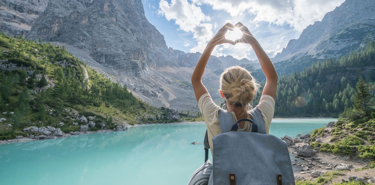 Frau von hinten fotografiert, steht vor einer weitläufigen Seenlandschaft in Italien und fängt mit ihrer zum Herz geformten Hand die Sonne ein