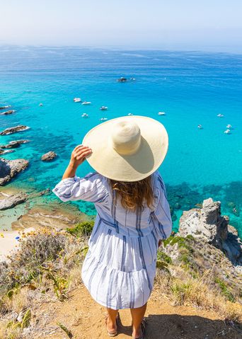 Eine Frau mit Hut steht auf Klippen und schaut auf das türkisfarbende Meer 