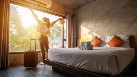 Frau steht mit Koffer im Hotelzimmer am Bett und blickt durch das große Fenster ins Licht und reißt die Arme hoch