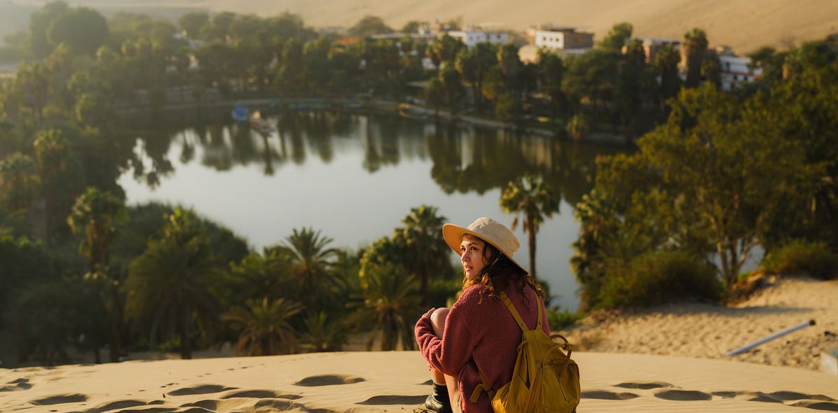 Frau sitzt im Sand und blickt auf die Oase Huacachina in Peru