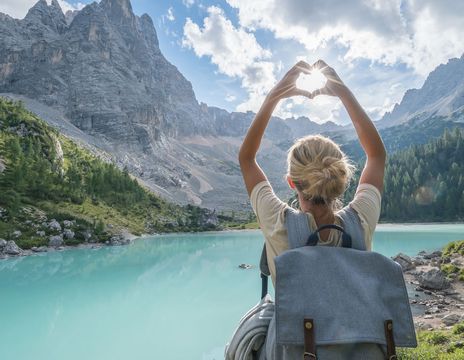 Frau von hinten fotografiert, steht vor einer weitläufigen Seenlandschaft in Italien und fängt mit ihrer zum Herz geformten Hand die Sonne ein