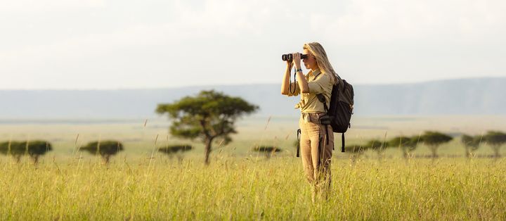 Frau steht während einer Safari mit Fernglas in weiter Graslandschaft mit Akazienbäumen im Hintergrund