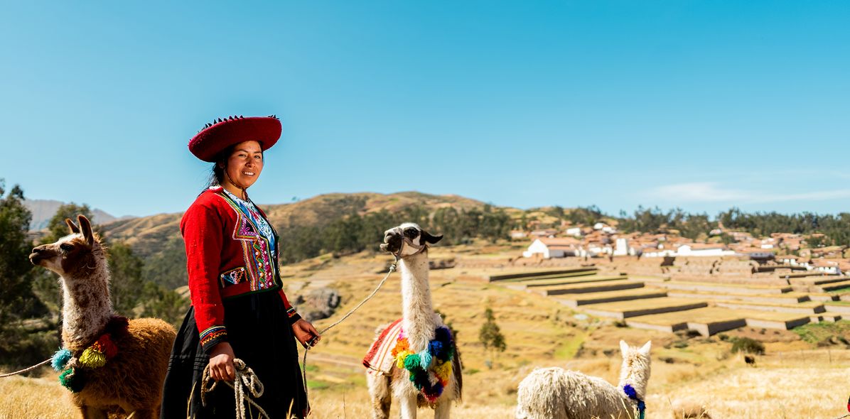 Frau in traditioneller Kleidung steht mit Alpakas auf einer Wiese in Peru