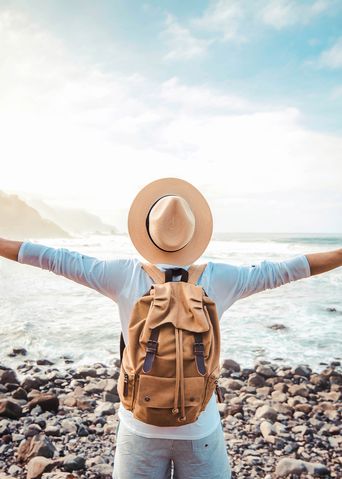 Mann mit Hut steht mit ausgebreiteten Armen auf felsigem Strand vor dem Meer