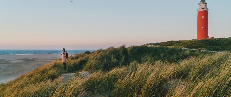 Frau steht in einer Dünenlandschaft vor einem Leuchtturm auf der Insel Texel