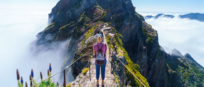 Eine Wanderin folgt einem schmalen Grat zwischen Felsen über dem Wolkenmeer Madeiras
