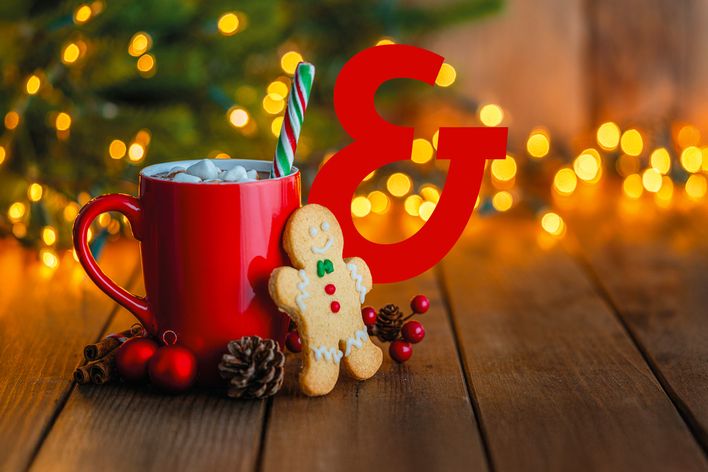 Ein Lebkuchenmännchen steht vor einer roten Weihnachtstasse mit Kakao vor einem beleuchtetem Weihnachtsbaum mit Berge&Meer Superzeichen