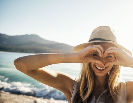 Frau mit Sommerhut am Strand formt mit den Händen ein Herz und schaut hindurch