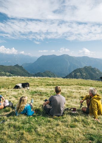 Wandergruppe genießt Rast auf grüner Almwiese mit Bergpanorama im Hintergrund