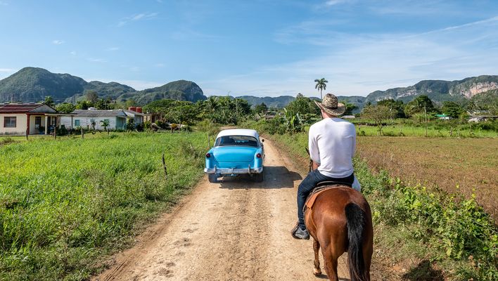 Reiter auf Pferd hinter einem typischen kubanischem Oldtimer auf einem Feldweg im Viñales-Tal auf Kuba
