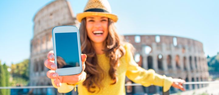 Frau mit Hut posiert vor dem Kolosseum in Rom und hält ihr Smartphone hoch