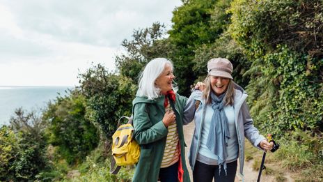 Zwei Freundinnen wandern lachend mit Rucksäcken auf sonnigem Waldweg