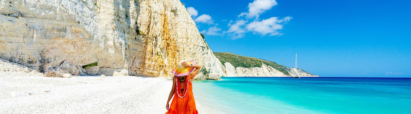 Frau von hinten spaziert auf weißem Sandstrand von Argostoli am türkisblauem Meer mit Felsen umrahmt in Kefalonia in Griechenland