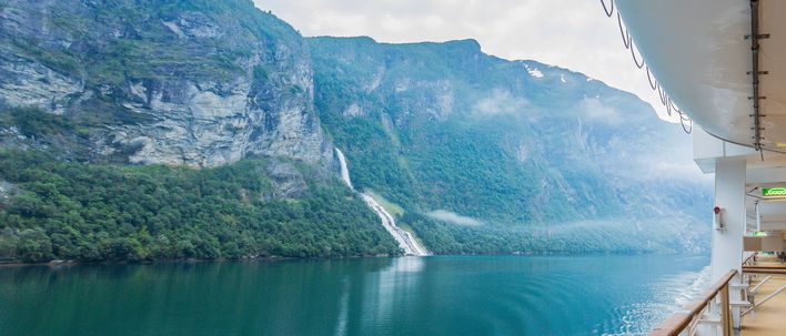 Blick vom Außendeck eines Kreuzfahrtschiffs auf einen Fjord mit steilen, grünen Berghängen und Wasserfall