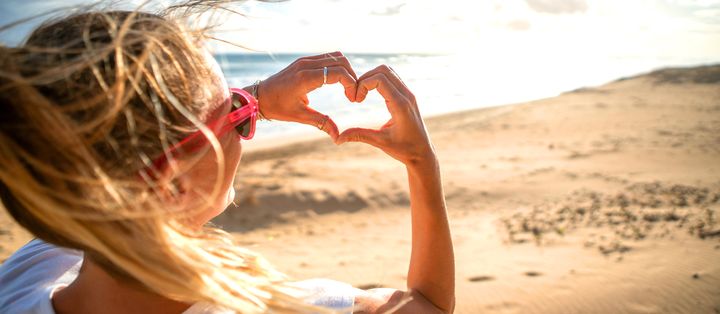 Frau in Rückansicht formt mit den Händen ein Herz am windigen Strand und blickt aufs Meer