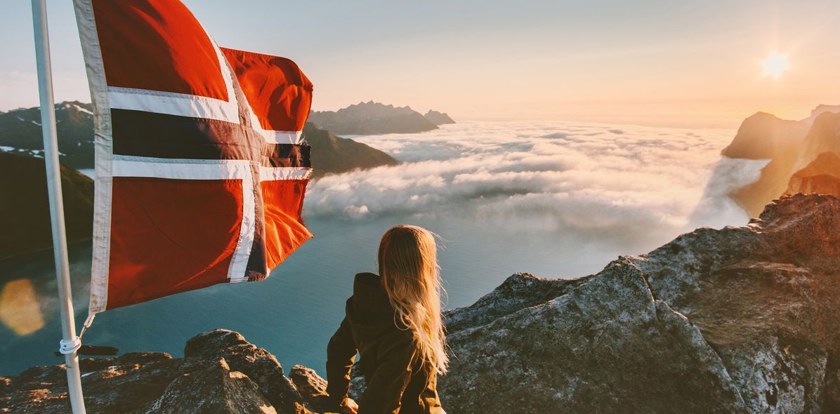 Eine Frau schaut auf ein nebliges Tal in Norwegen herab mit der norwegischen Flagge die neben ihr im Wind weht