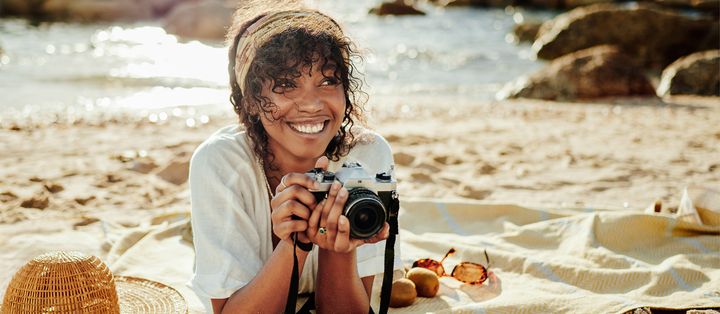 Frau mit Kopftuch und Kamera in der Hand liegt am Strand vor dem Meer mit Felsen in Sepiafarbton