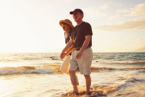 Best-Ager-Paar spaziert lachend bei Sonnenuntergang am Strand entlang