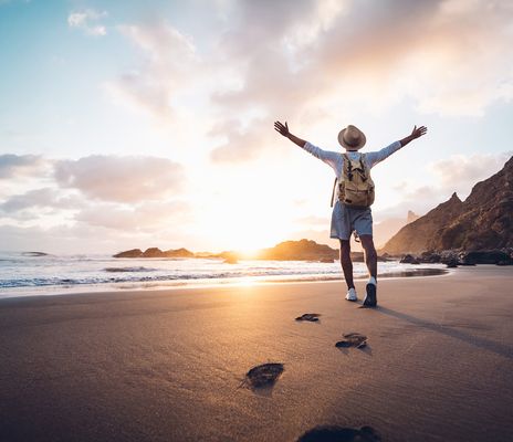 Reisender mit Rucksack genießt den Sonnenuntergang am Strand mit ausgebreiteten Armen