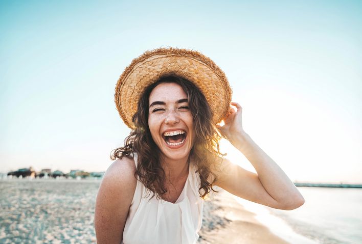Frau mit Sonnenhut steht am Strand und lacht in die Kamera