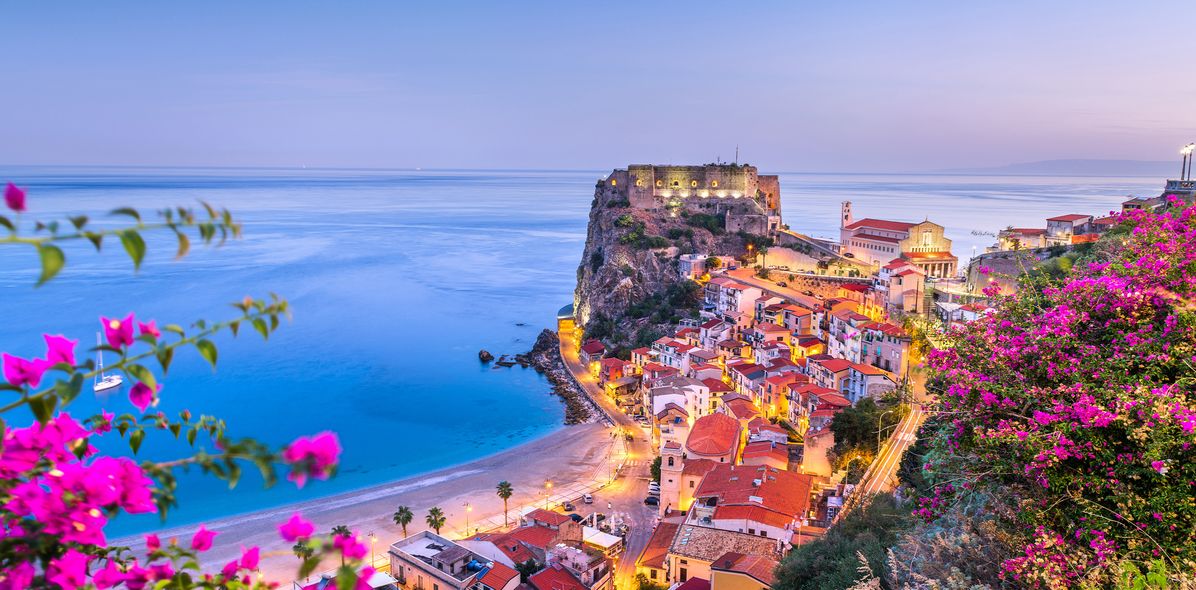 Panoramablick auf die Küste von Scilla in Kalabrien mit bunten Häusern am Hang, blauem Meer und pink blühenden Bougainvillea im Vordergrund