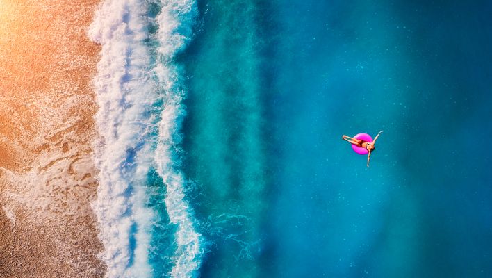 Frau treibt entspannt auf pinkem Schwimmring im türkisblauen Meer mit weißen Wellen am Strand von oben gesehen