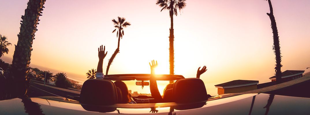 Zwei Frauen sitzen in einem Cabrio welches der Sonne entgegen und von Palmen umgeben auf einem Parkplatz steht und die Hände in die Höhe strecken