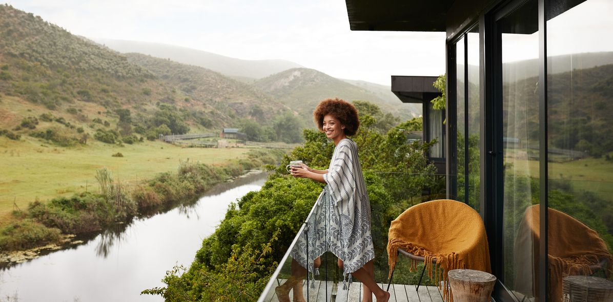 Eine Reisende genießt von der Terrasse einer Safari-Lodge den Blick auf einen Fluss und grüne Hügel
