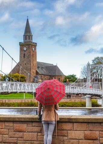 Frau mit rotem Regenschirm steht am Fluss Ness mit Blick auf die Brücke der Stadt Inverness