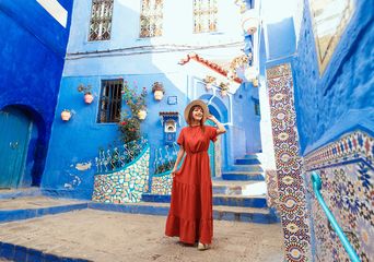 Eine Frau im Roten Kleid mit Hut spaziert durch die blauen Gassen von Chefchaouen
